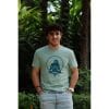 Hombre sonriendo con Camiseta Positano de Flow Mico en verde salvia con manos en bolsillos en entorno natural